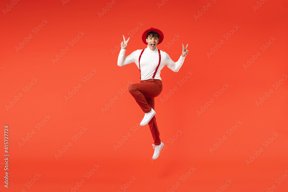 Full length of young spanish latinos overjoyed smiling fun happy man in hat white shirt trousers with suspenders jumping high show v-sign victory gesture isolated on red background studio portrait.