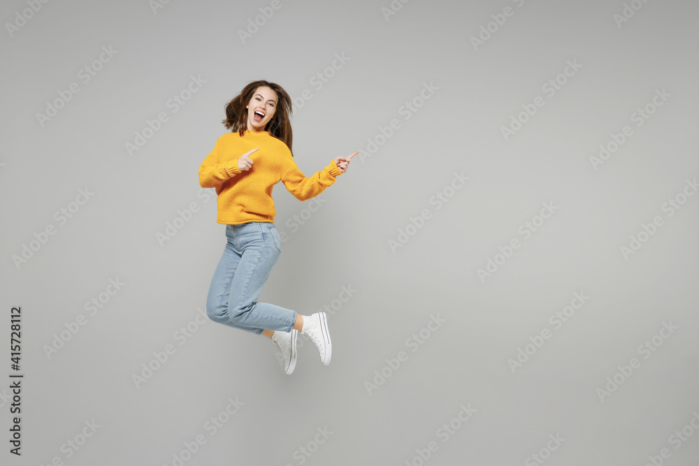 Full length of young excited overjoyed expressive woman in knitted yellow sweater run fast in air jump high point index finger aside on workspace mock up isolated on grey background studio portrait.. Full length of young excited overjoyed expressive woman in knitted yellow sweater run fast in air jump high point index finger aside on workspace mock up isolated on grey background studio portrait..