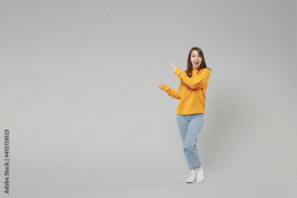 Full length of young smiling positive caucasian happy woman 20s in knitted yellow sweater stand point index finger aside on workspace copy space area isolated on grey color background studio portrait - obrazy, fototapety, plakaty 