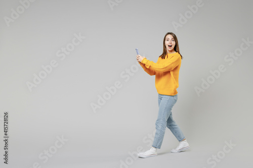 Full length of young surprised shocked caucasian happy woman 20s wear knitted yellow sweater going walking going hold mobile cell phone look camera isolated on grey color background studio portrait © ViDi Studio