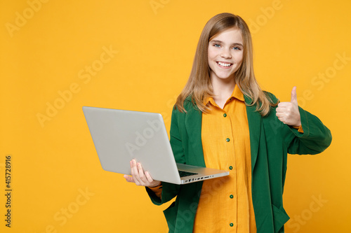 Little blonde pretty cute kid girl 12-13 years old in casual clothes hold laptop pc computer show thumb up gesture isolated on yellow background children studio portrait. Childhood lifestyle concept