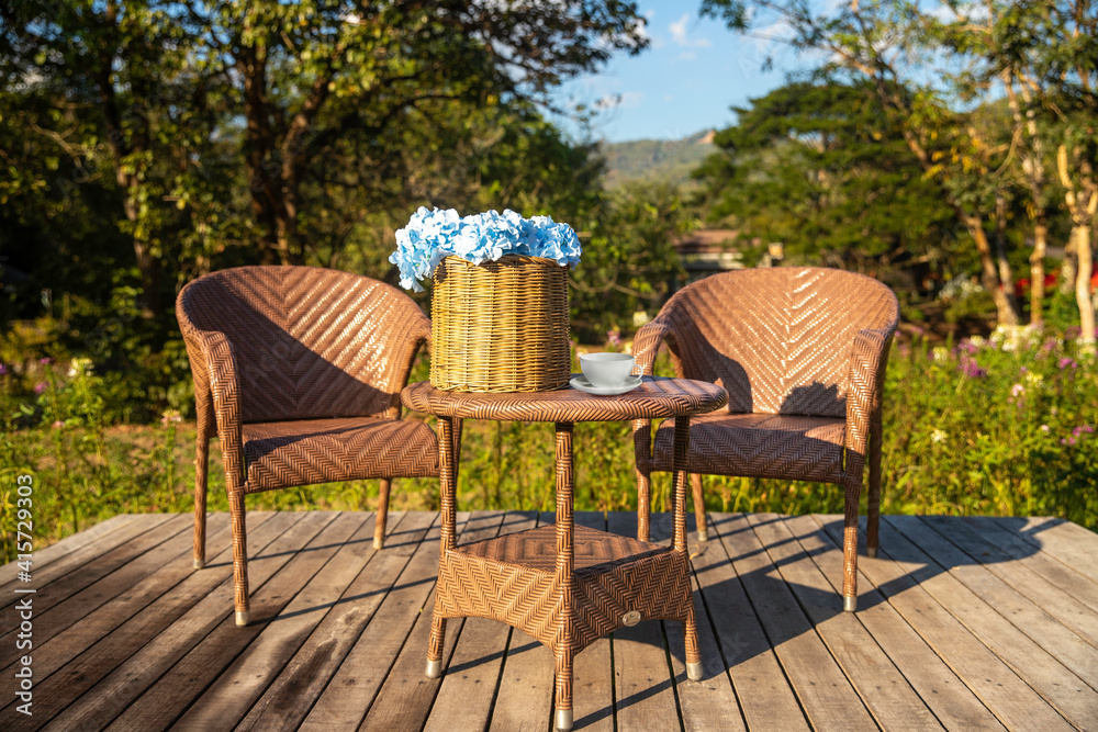 Fototapeta premium Two armchairs, wooden garden furniture on wood floor outdoor for relaxing on summer days. Garden landscape with two chairs in nature, flowers in vase and coffee cup..