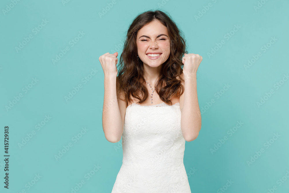 Happy funny bride young woman in white wedding dress doing winner gesture celebrating clenching fists say yes isolated on blue turquoise background studio portrait. Ceremony celebration party concept.