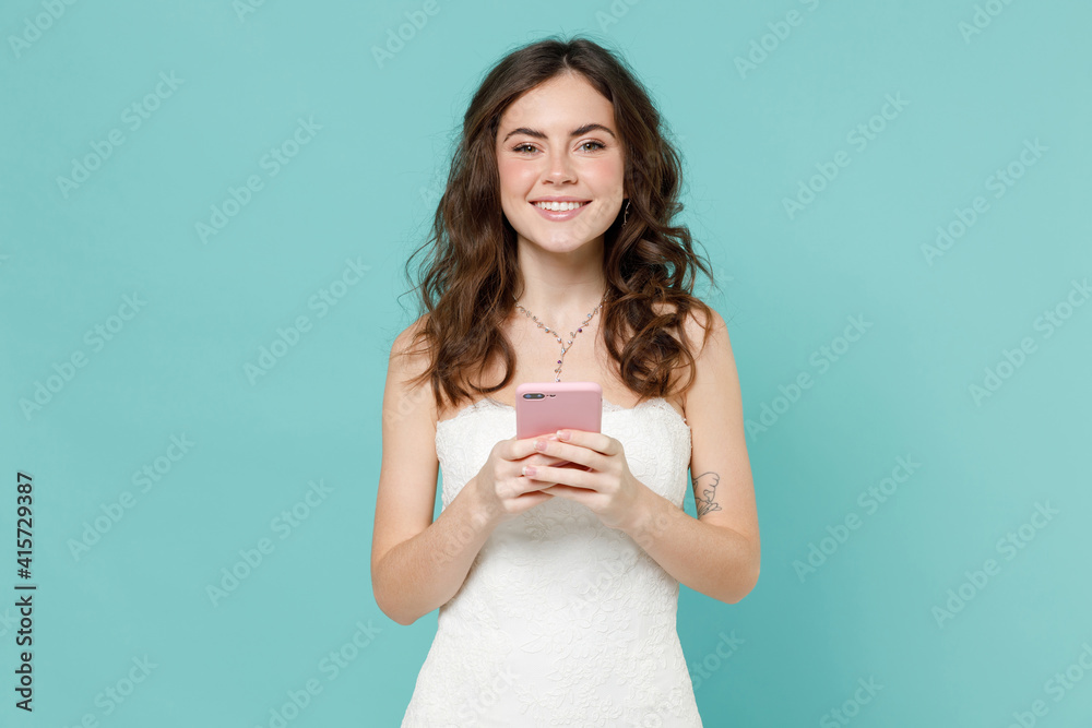 Cheerful bride young woman 20s in beautiful white wedding dress using mobile cell phone typing sms message isolated on blue turquoise background studio portrait. Ceremony celebration party concept. Cheerful bride young woman 20s in beautiful white wedding dress using mobile cell phone typing sms message isolated on blue turquoise background studio portrait. Ceremony celebration party concept.