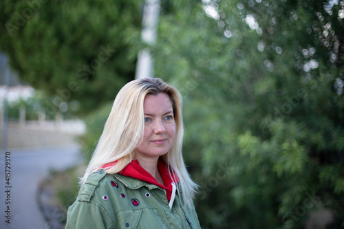 Closeup portrait of a beautiful forty-year-old blonde caucasian woman in a casual green jacket posing outdoors in a park against the background of green trees