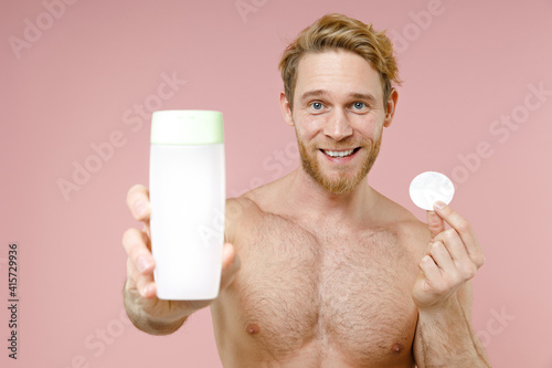 Cheerful bearded naked young man 20s perfect skin hold bottle of micelar water lotion tonic cotton pad isolated on pink background studio portrait. Skin care healthcare cosmetic procedures concept. photo