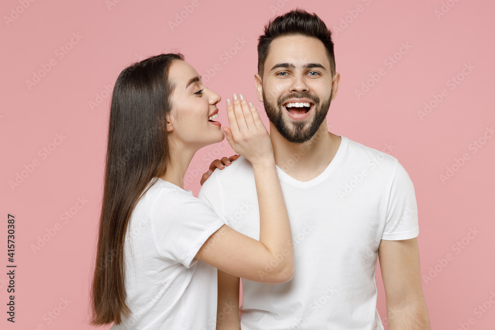 Young couple two friends bearded man brunette woman in white basic blank print design t-shirt whispering gossip and tells secret behind her hand, sharing news isolated on pastel pink color background Young couple two friends bearded man brunette woman in white basic blank print design t-shirt whispering gossip and tells secret behind her hand, sharing news isolated on pastel pink color background