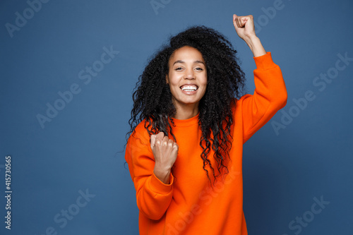 Cheerful joyful young african american woman in casual basic orange sweatshirt doing winner gesture celebrating clenching fists say yes looking camera isolated on blue wall background studio portrait.