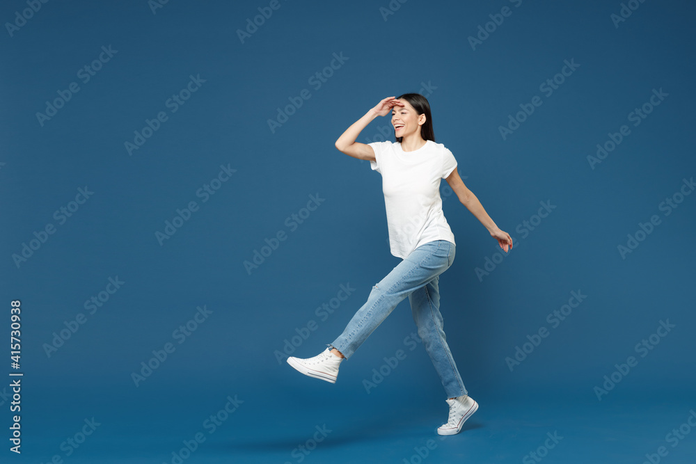 Full length young smiling latin woman 20s wearing white casual basic t-shirt holding hand at forehead looking far away distance raised up leg for step isolated on dark blue background studio portrait.