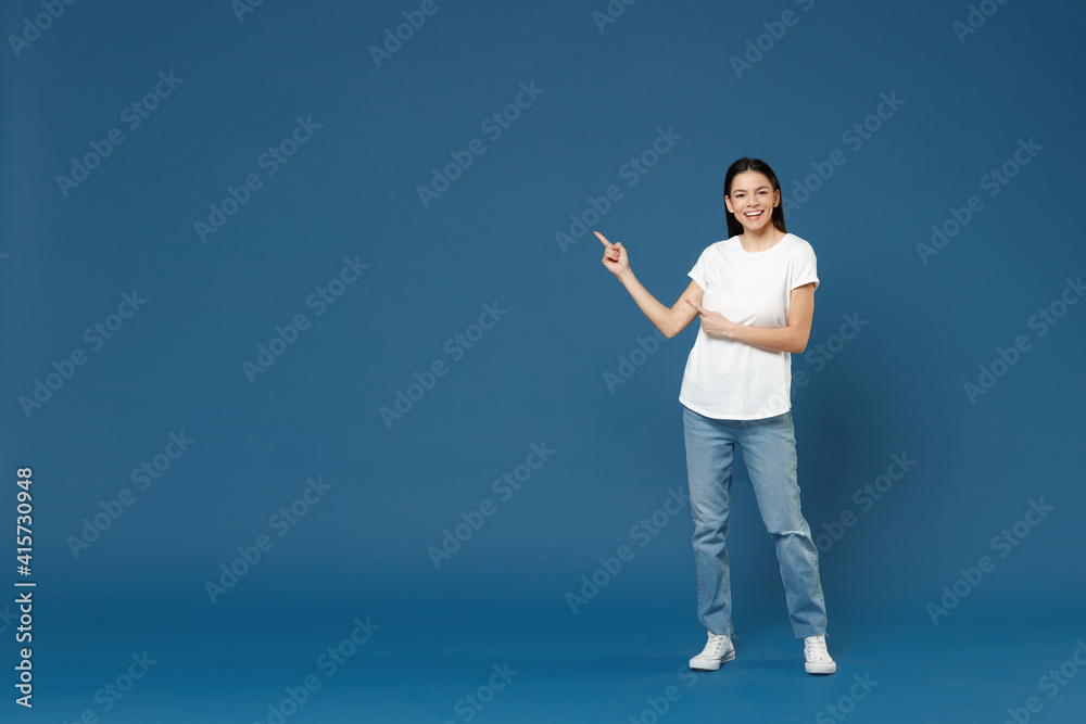 Full length young smiling latin woman 20s wearing white casual basic t-shirt pointing index finger aside back on copy space mock up area workspace isolated on dark blue background studio portrait. Full length young smiling latin woman 20s wearing white casual basic t-shirt pointing index finger aside back on copy space mock up area workspace isolated on dark blue background studio portrait.