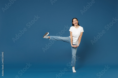 Full length of young smiling relaxed beautiful latin woman 20s in white basic t-shirt keep hands outstretched with raised up leg fingers intertwined isolated on dark blue background studio portrait.