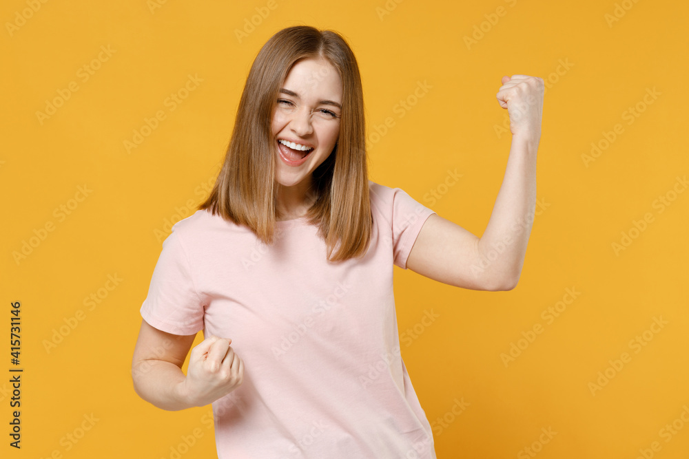 Young overjoyed happy woman 20s with nude make up wearing casual basic pastel pink t-shirt, blank print design do winner gesture clench fist celebrating isolated on yellow background studio portrait