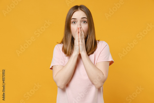Young frightened shocked impressed woman 20s with nude make up wearing casual basic pastel pink t-shirt covering face with hands looking camera isolated on yellow color background studio portrait