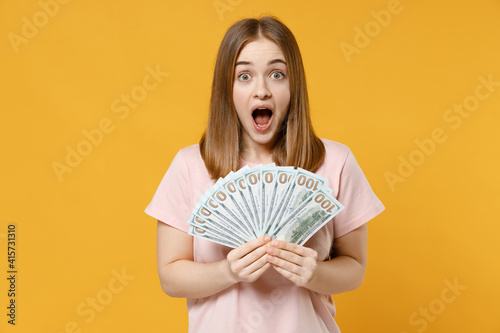Obraz na plátně Young overjoyed surprised shocked rich student woman 20s wearing casual basic pastel pink t-shirt hold fan of cash money in dollar banknotes look camera isolated on yellow background studio portrait