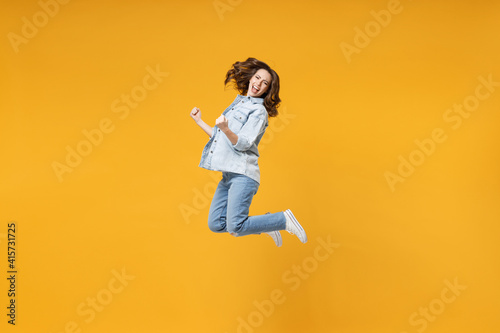 Full length of young overjoyed excited fun student happy woman 20s in denim shirt white t-shirt do winner gesture clench fist celebrating jump high isolated on yellow color background studio portrait