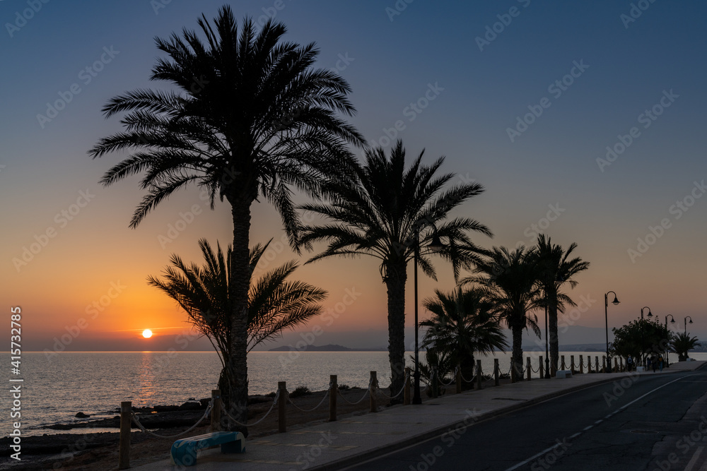 sunset over the ocean with palm trees in silhouette and a beachfront sidewalk and oceanfront road