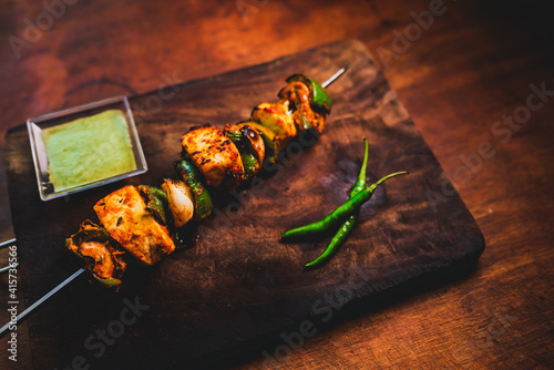 Obraz na plátně Spicy Indian paneer tikka coated in red sauce and flame grilled on skewer and served on a dark wooden plate with green chilli and coriander sauce