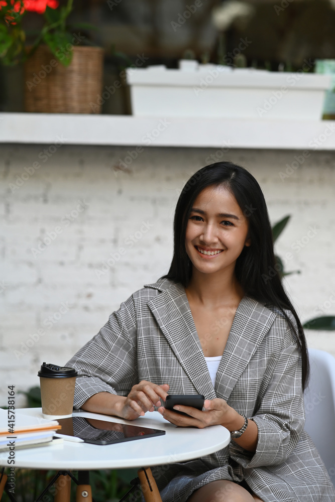 © Prathankarnpap - Attractive businesswoman holding smart phone and smiling to camera while sitting in cafe .