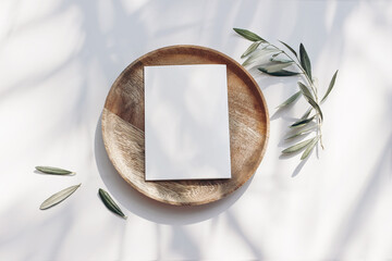 Summer wedding stationery mock-up scene. Blank greeting card, wooden plate, olive tree leaves and branches in sunlight. White table background with palm shadows. Feminine flat lay, top view.