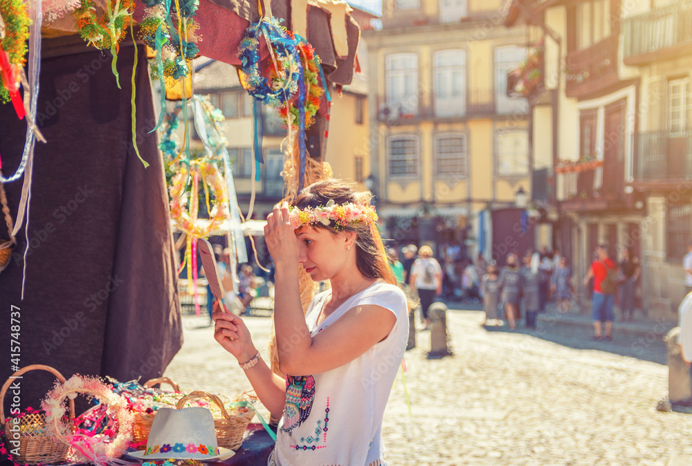 Young woman traveler trying on flower crown wreath and looking at hand mirror near souvenir booth stand during city day in Guimaraes medieval old historical town centre, Portugal