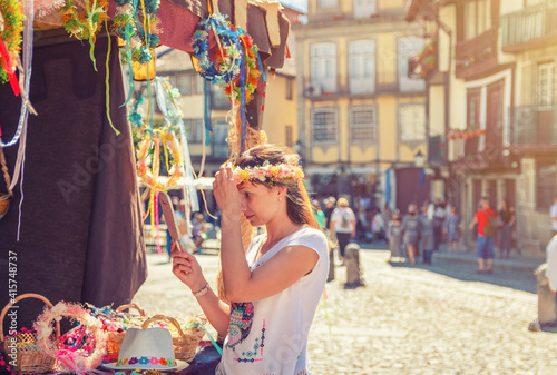 Young woman traveler trying on flower crown wreath and looking at hand mirror near souvenir booth stand during city day in Guimaraes medieval old historical town centre, Portugal © Aliaksandr