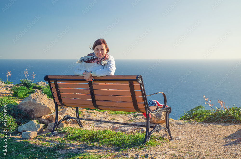 Young beautiful girl traveler with white jacket looking at camera, sit and smile on bench at Dingli Cliffs with Mediterranean sea blue water background, tourist woman on vacation in Malta island