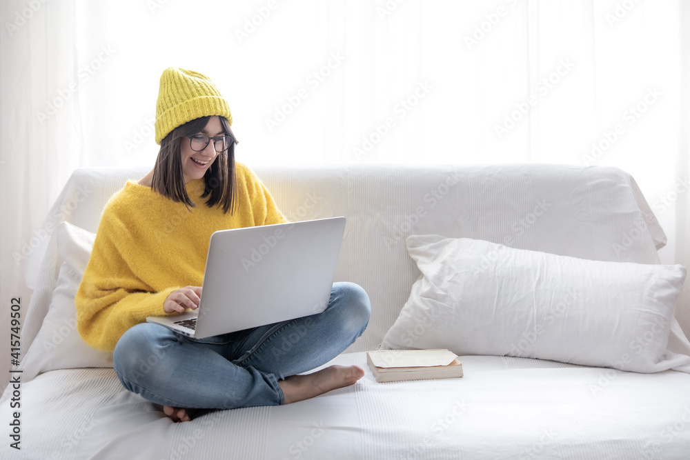 A young girl in a yellow sweater and yellow hat sits on the couch with a laptop and smiles.