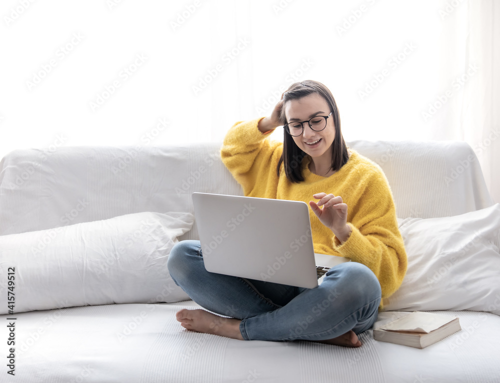 A cute girl in a yellow sweater and glasses sits with a laptop at home on the couch and smiles. A cute girl in a yellow sweater and glasses sits with a laptop at home on the couch and smiles.