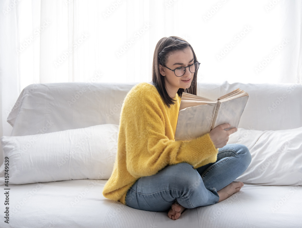A cute girl in glasses and a yellow sweater is sitting on the sofa with a book.
