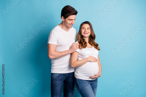 Photo of cheerful sweet cute couple expecting child hugging belly looking each other isolated blue color background © deagreez