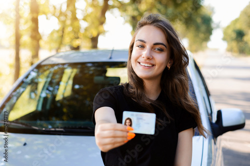 Pretty girl with cheerful smile standing near the car and showing driving license to the camera. Woman expresses her happiness after passing the driving exam.