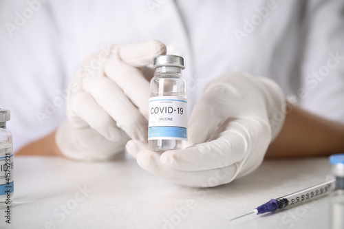 Doctor holding vial with vaccine against Covid-19 at table, closeup