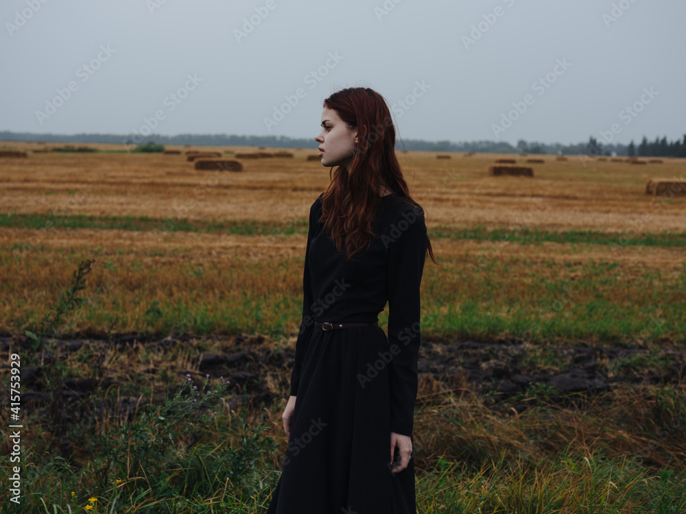 Obraz premium Beautiful woman in a black dress with red hair and a dry field in the background