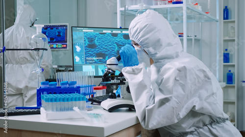 Chemist in protection suit typing on pc and analyzing virus sample using microscope in scientific lab. Team of doctors examining vaccine evolution using high tech researching diagnosis against covid19