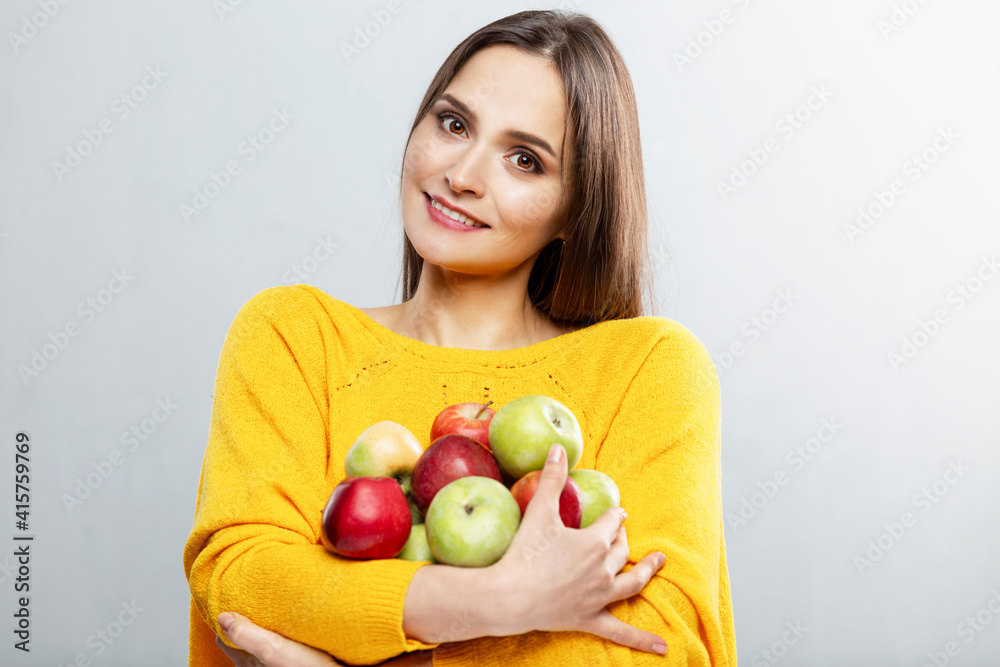 Young smiling woman holding many red and green apples in her hands. Beautiful brunette in a yellow sweater. Healthy food, vitamins and vegetarianism. Grey background. Young smiling woman holding many red and green apples in her hands. Beautiful brunette in a yellow sweater. Healthy food, vitamins and vegetarianism. Grey background.