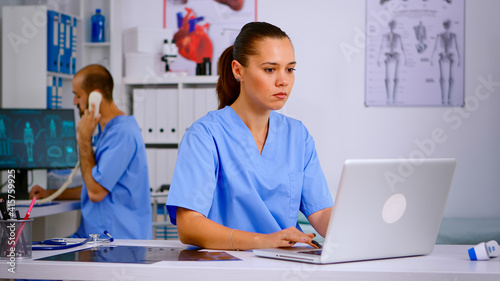 Medical practitioner typing patient health report on laptop in hospital office. Health care staff sitting at desk talking at phone in background, using computer in clinic looking at monitor, medicine.