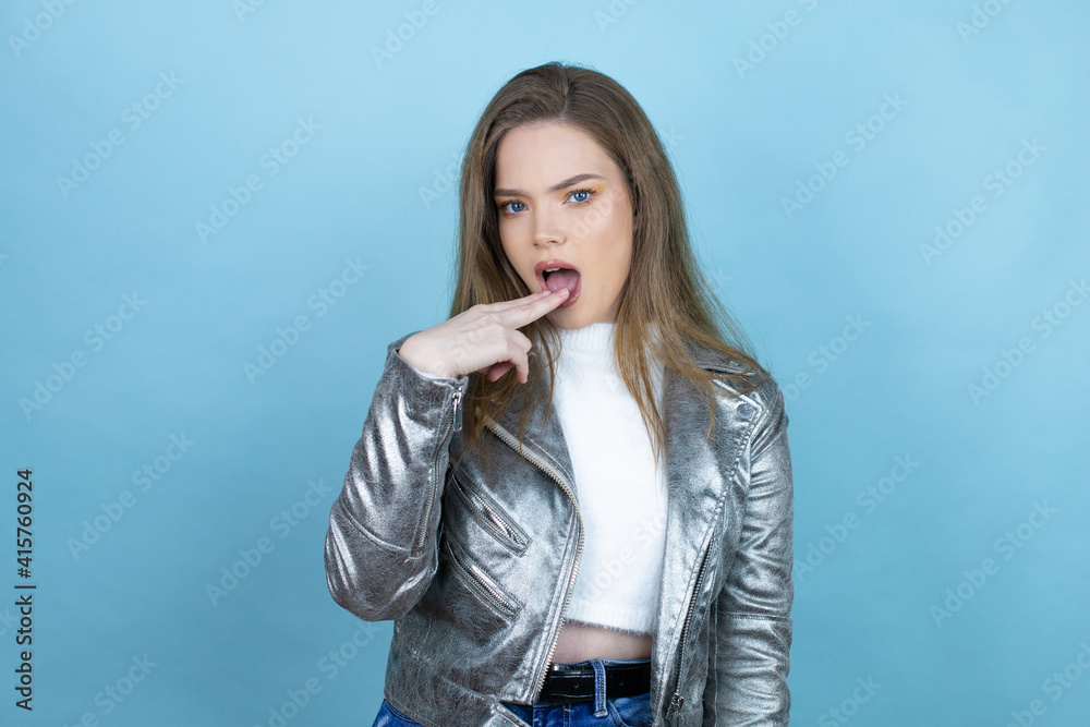 Pretty woman with long hair wearing a casual jacket over blue background disgusted with her hand inside her mouth
