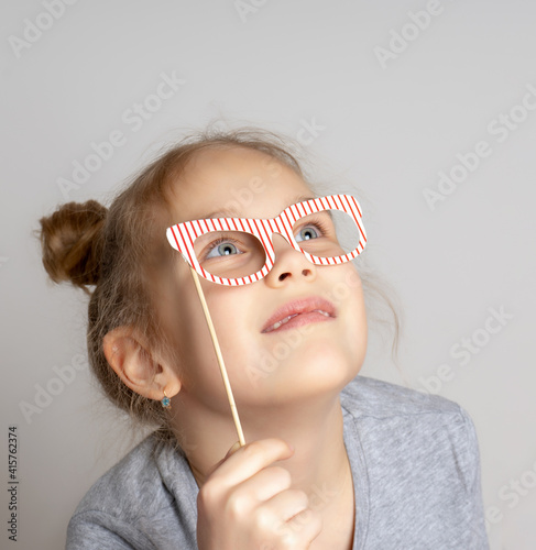 Pensive little girl in carnival mask with paper glasses. Festive costume for a masquerade. Attractive female child posing with photo booth accessory studio portrait shot