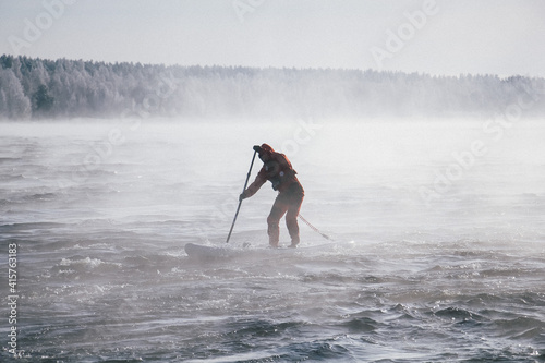 Surfer in a red wetsuit on a board with a paddle floats against the waves. Winter sup surfing on the Vuoksa river. Losev rapids. photo