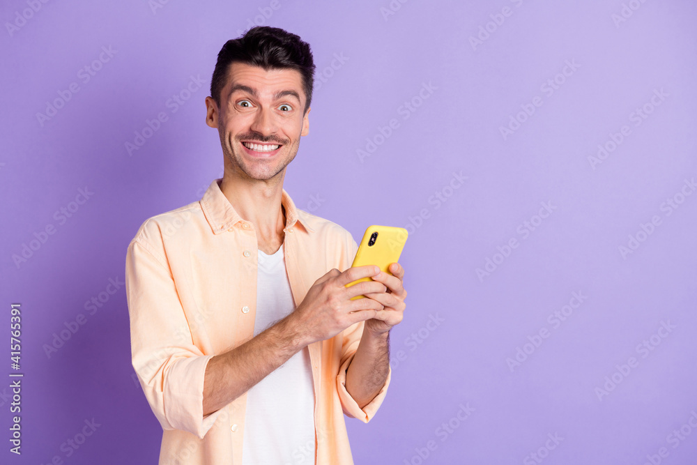 Photo of optimistic brunet guy hold telephone wear peach shirt isolated on lilac color background Photo of optimistic brunet guy hold telephone wear peach shirt isolated on lilac color background