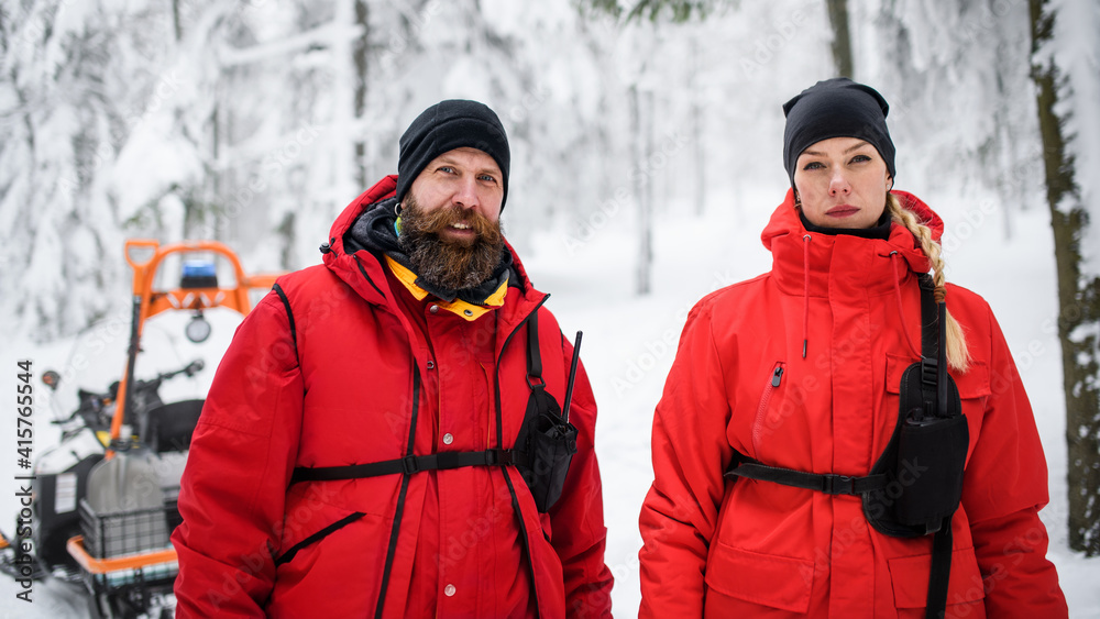 Woman and man paramedic from mountain rescue service outdoors in winter in forest, looking at camera. Woman and man paramedic from mountain rescue service outdoors in winter in forest, looking at camera.