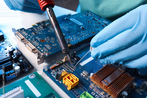 Technician repairing electronic circuit board with soldering iron at table, closeup photo