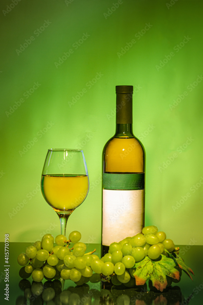 A green bottle of white wine next to a glass filled with wine, a bunch of grapes in front. on a green background