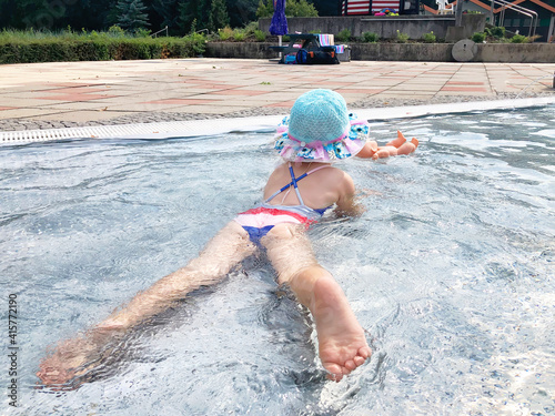 Little toddler girl splashing in an outdoors swimming pool on warm summer day. Happy healthy child enjoying sunny weather in city public pool.