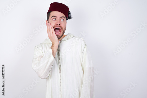 young handsome Caucasian man wearing Arab djellaba and Fez hat over white wall excited looking to the side hand on face. Advertisement and amazement concept.