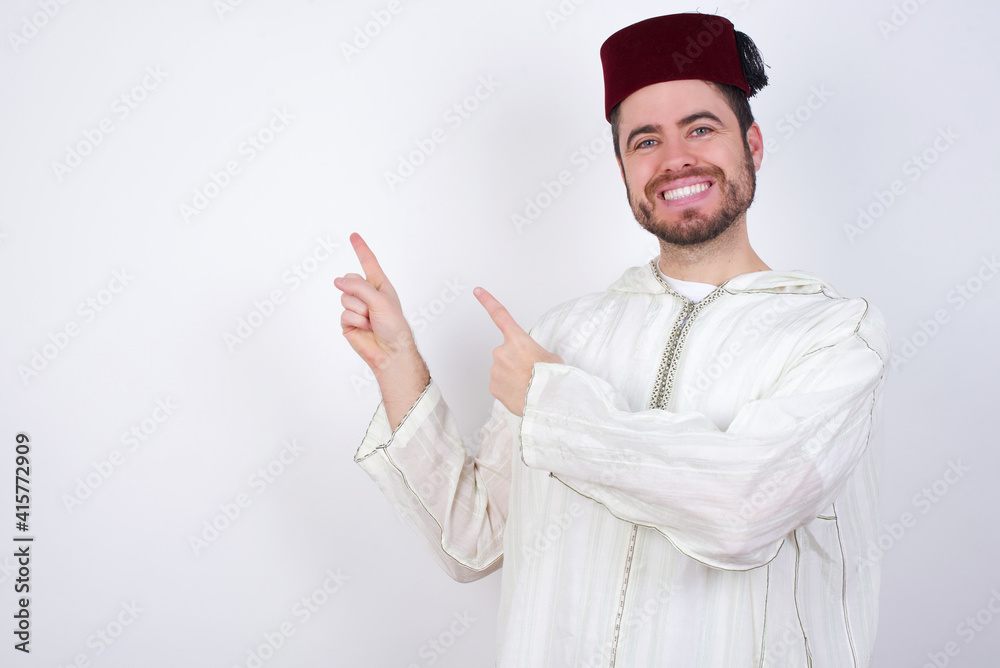 young handsome Caucasian man wearing Arab djellaba and Fez hat over white wall with positive expression, indicates with fore finger at blank copy space for your promotional text or advertisement. young handsome Caucasian man wearing Arab djellaba and Fez hat over white wall with positive expression, indicates with fore finger at blank copy space for your promotional text or advertisement.