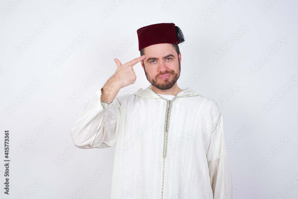 Unhappy young handsome Caucasian man wearing Arab djellaba and Fez hat over white wall curves lips and makes suicide gesture, shoots in temple with hand, tries kill himself. Unhappy young handsome Caucasian man wearing Arab djellaba and Fez hat over white wall curves lips and makes suicide gesture, shoots in temple with hand, tries kill himself.
