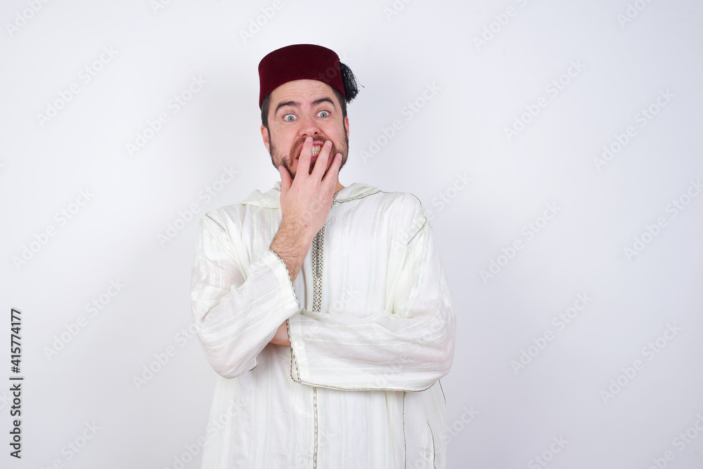 young handsome Caucasian man wearing Arab djellaba and Fez hat over white wall covering mouth with hands scared from something or someone bitting nails young handsome Caucasian man wearing Arab djellaba and Fez hat over white wall covering mouth with hands scared from something or someone bitting nails