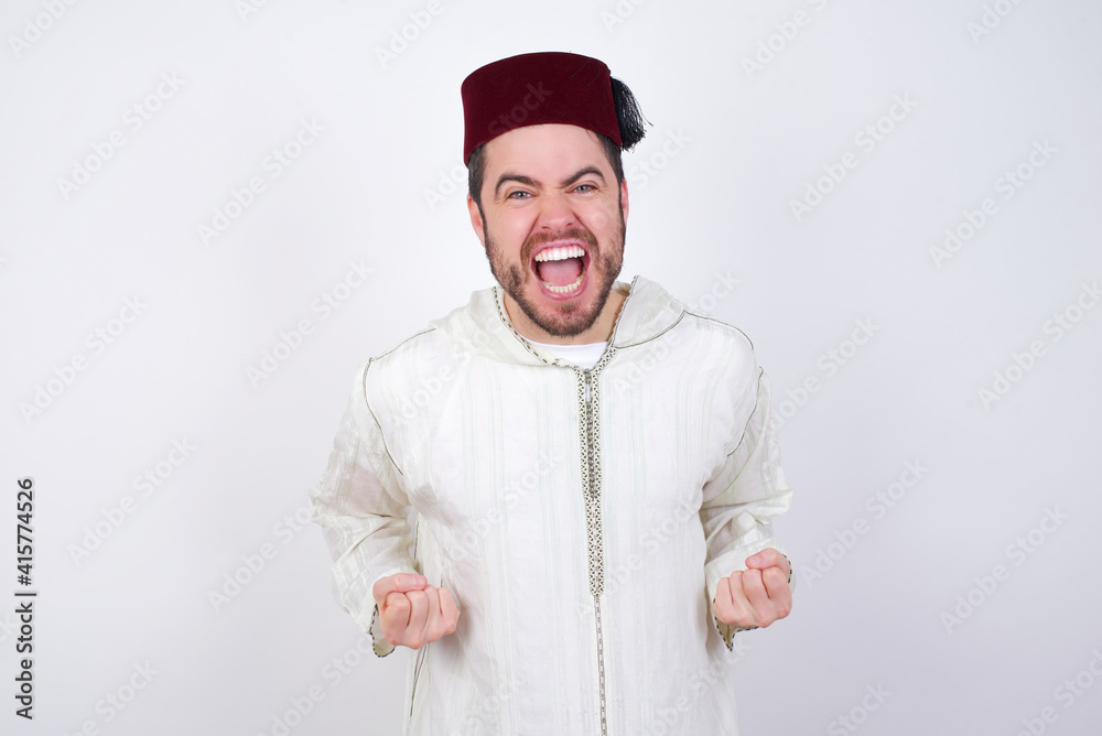 young handsome Caucasian man wearing Arab djellaba and Fez hat over white wall raising fists up screaming with joy being happy to achieve goals. young handsome Caucasian man wearing Arab djellaba and Fez hat over white wall raising fists up screaming with joy being happy to achieve goals.