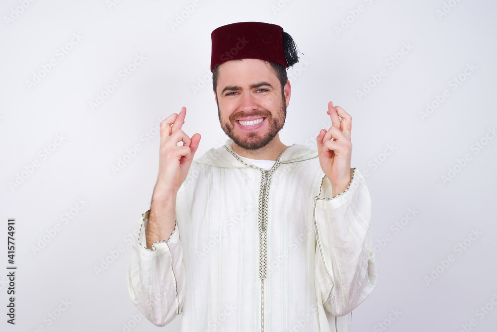 young handsome Caucasian man wearing Arab djellaba and Fez hat over white wall holding fingers crossed with worried expression hoping boss didn't noticed mistakes at work. young handsome Caucasian man wearing Arab djellaba and Fez hat over white wall holding fingers crossed with worried expression hoping boss didn't noticed mistakes at work.